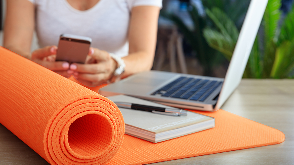 Woman sat at a desk with a laptop, mobile phone, notepad and yoga mat
