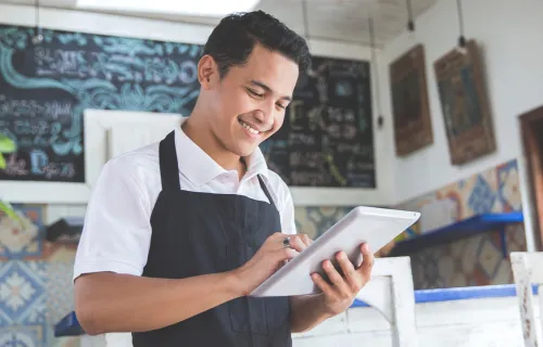 Male café owner wearing a black apron smiling while using a tablet